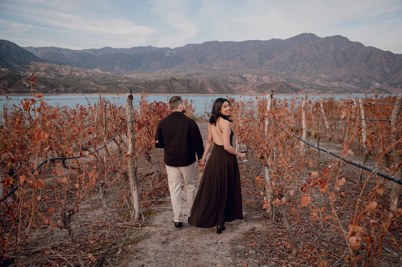 Pareja caminando de la mano entre viñedos otoñales, con montañas y un lago al fondo